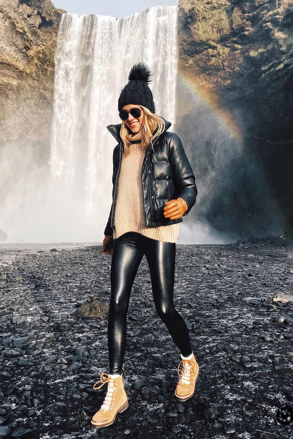 image of a woman walking in front of a waterfall wearing faux leather leggings, brown shearling boots, a sweater, puffer jacket, and toque