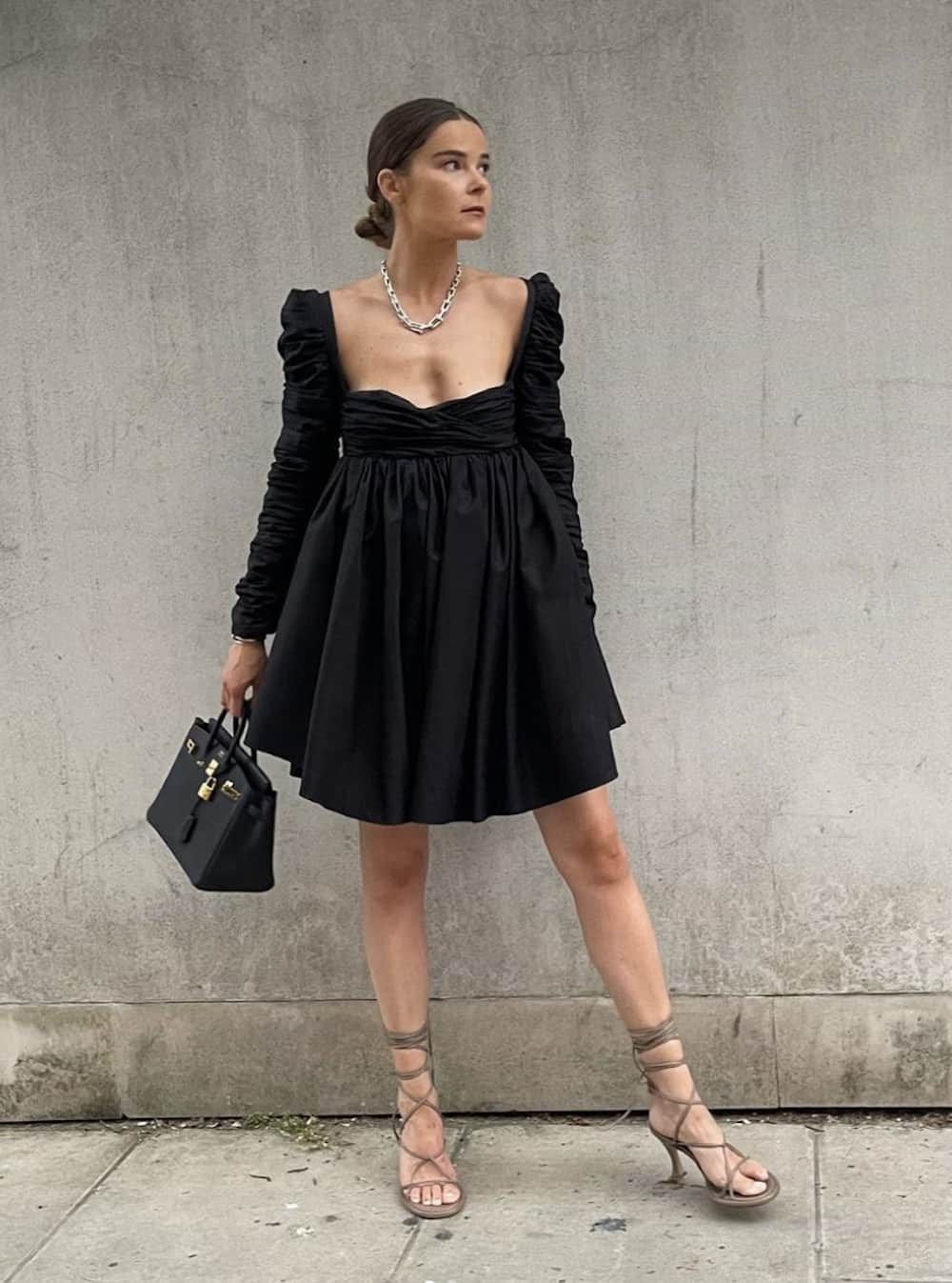 image of a woman in a black mini dress with long sleeves and strappy flat sandals holding a small designer black bag