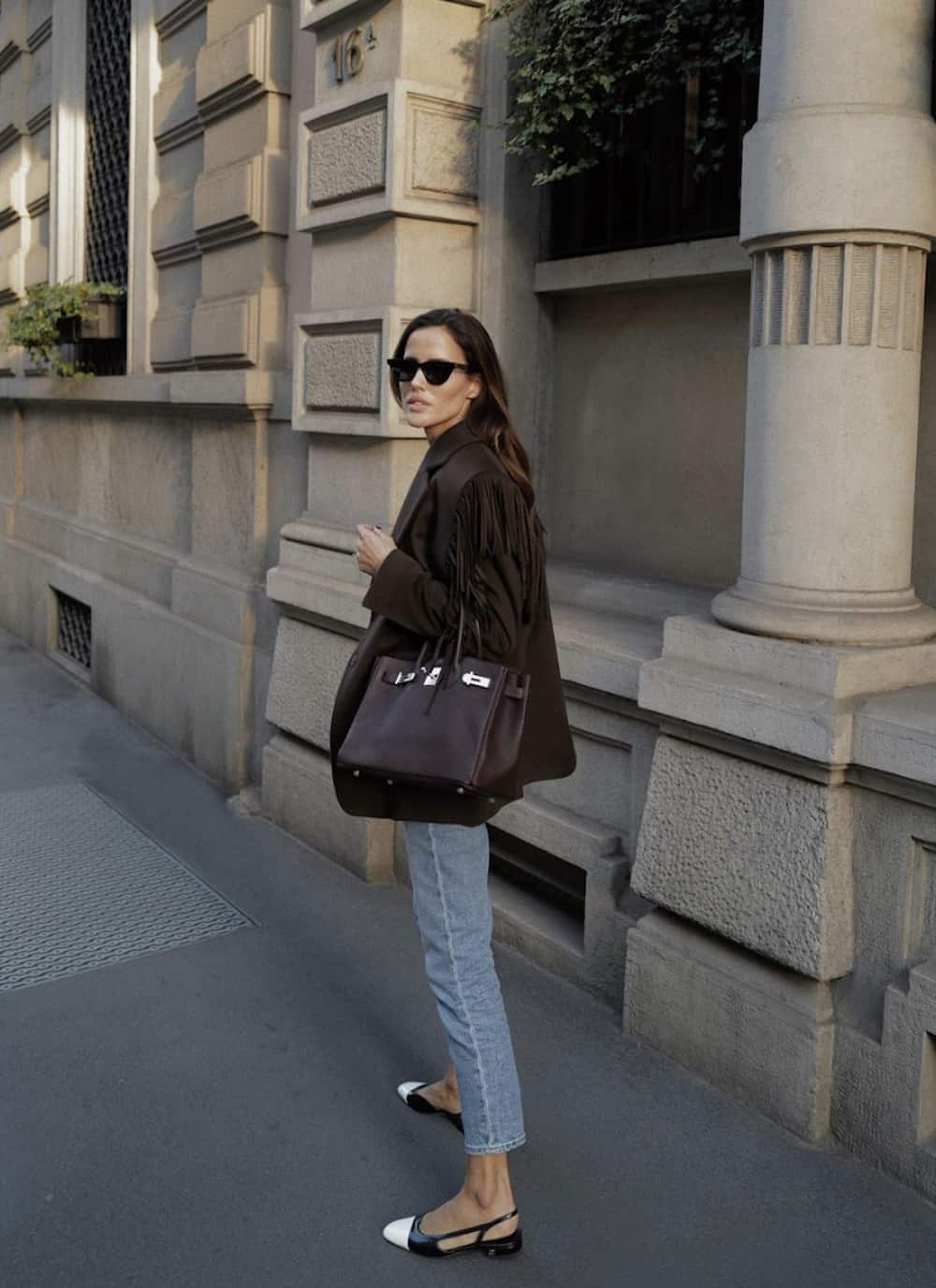 image of a woman in a brown fringe jacket, with a brown Birkin bag, wearing blue jeans, and cap toe flats