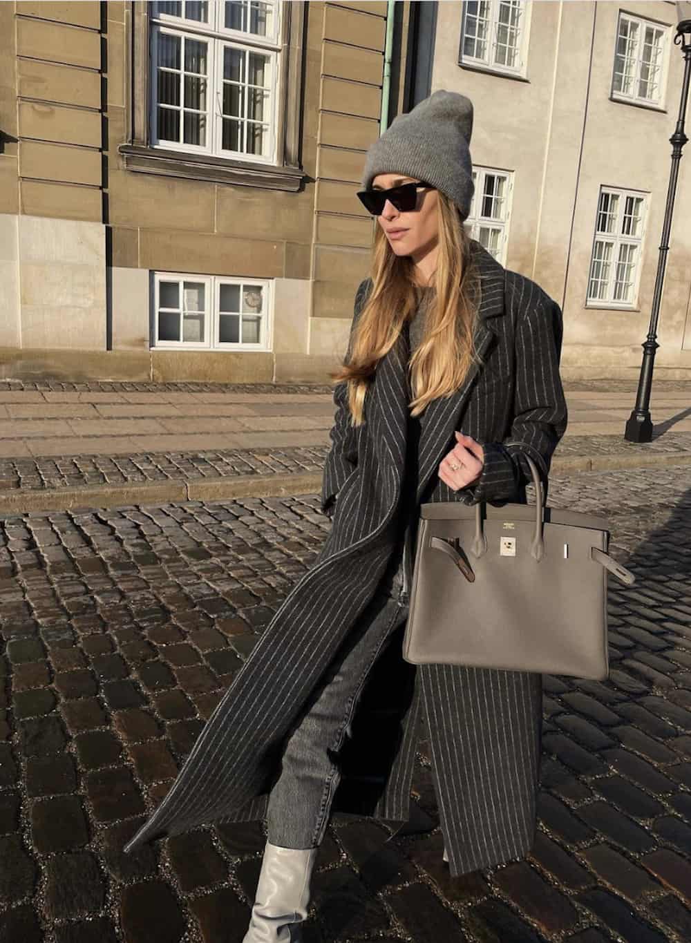 image of a woman in a wool trench coat and grey toque carrying a Hermes Birkin bag in grey