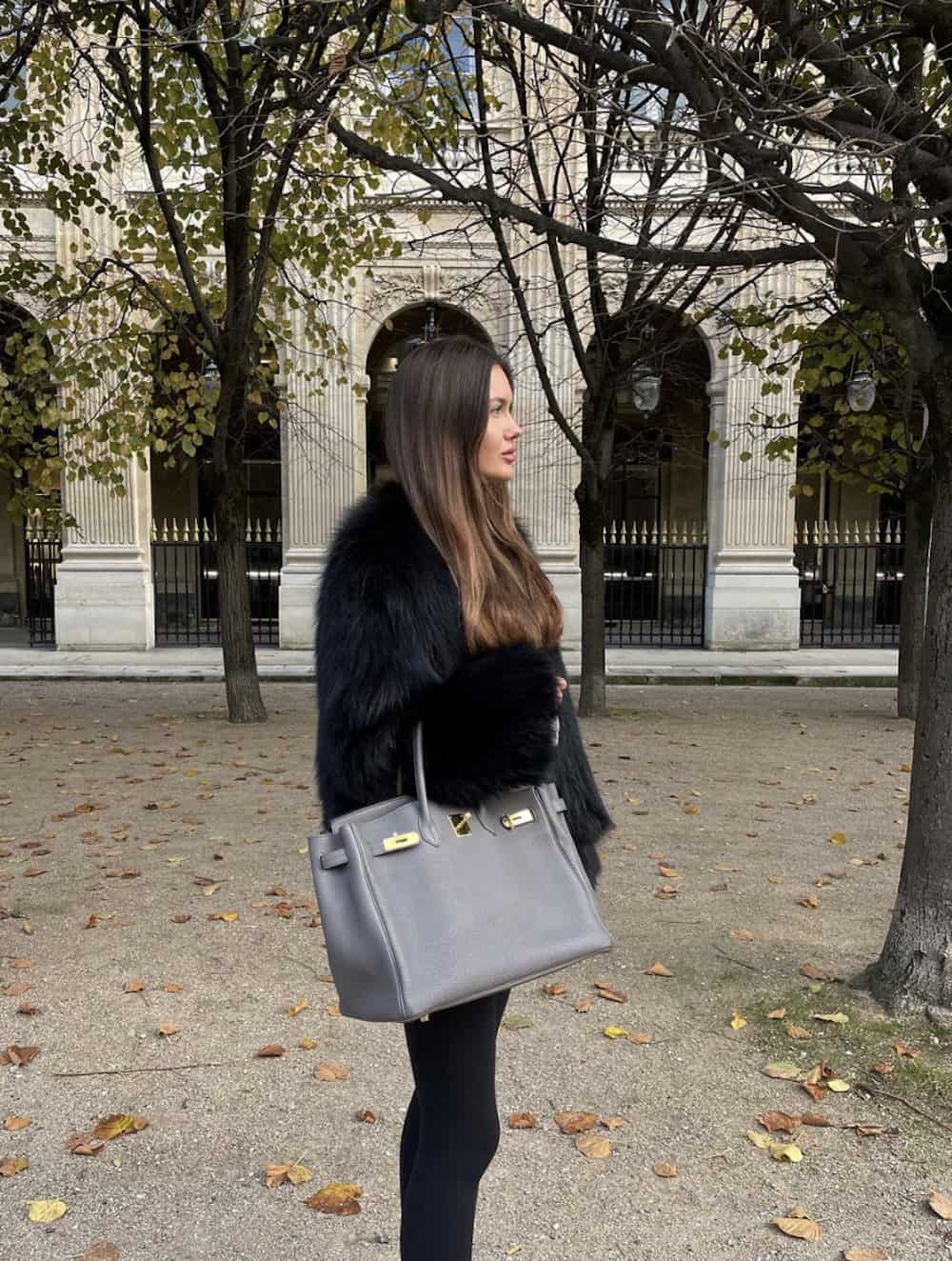 image of a woman in a black faux fur coat carrying a grey Hermes Birkin bag