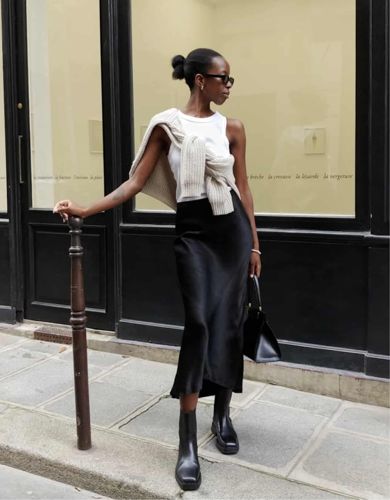 Woman wearing a black slip skirt, a white tank top, boots and a cream cardigan tied around her neck.