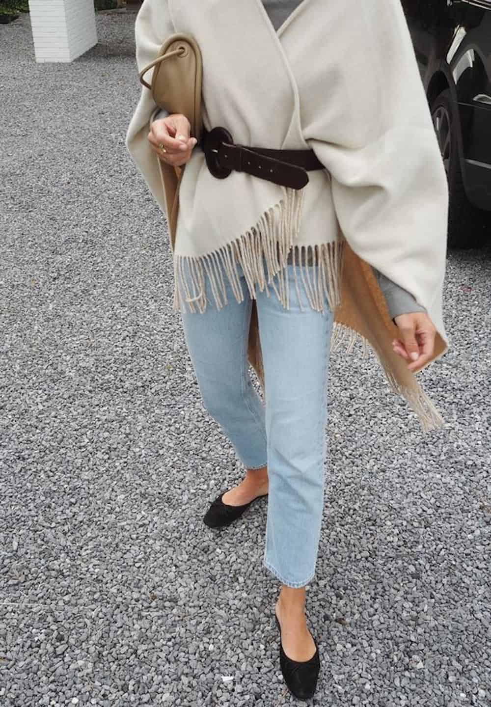 image of a woman from the shoulders down wearing a poncho with a belt, blue jeans, and black flats