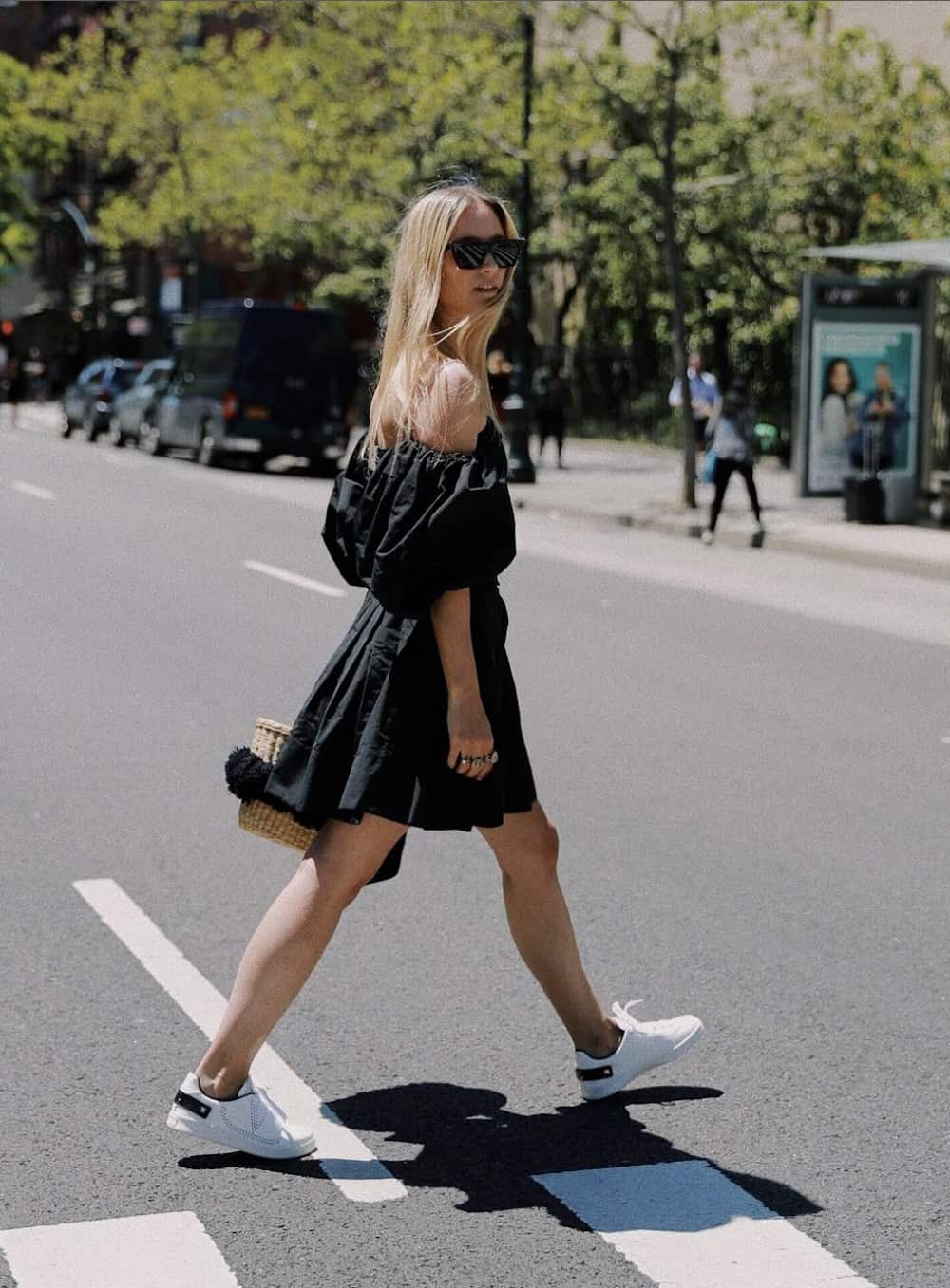 Woman wearing a black dress, sneakers and carrying a straw bag.