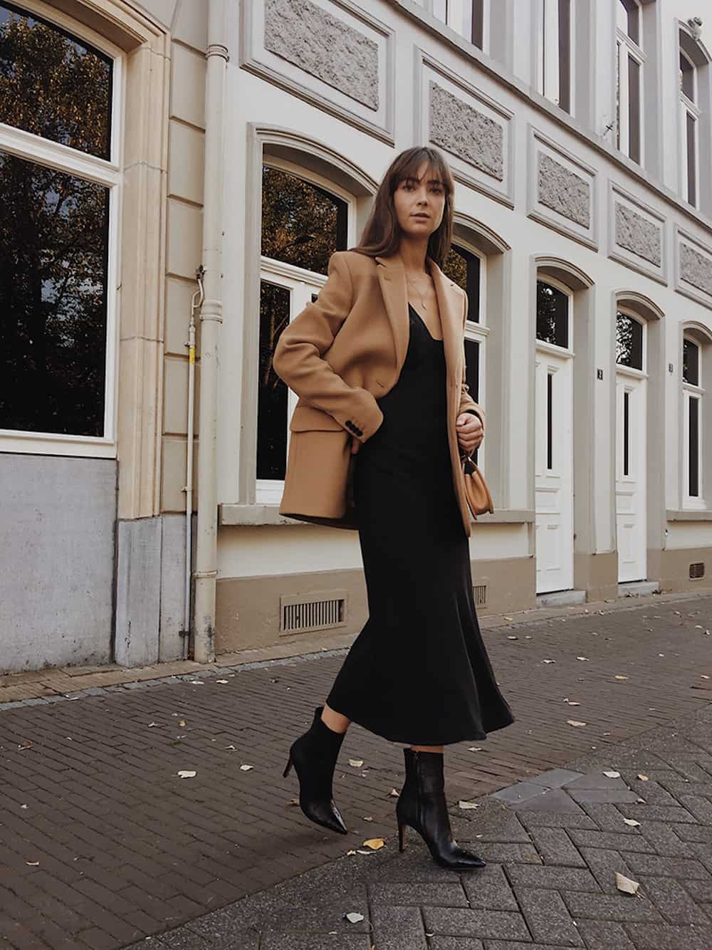 Woman wearing a black midi dress, black heeled booties and a camel colored coat.