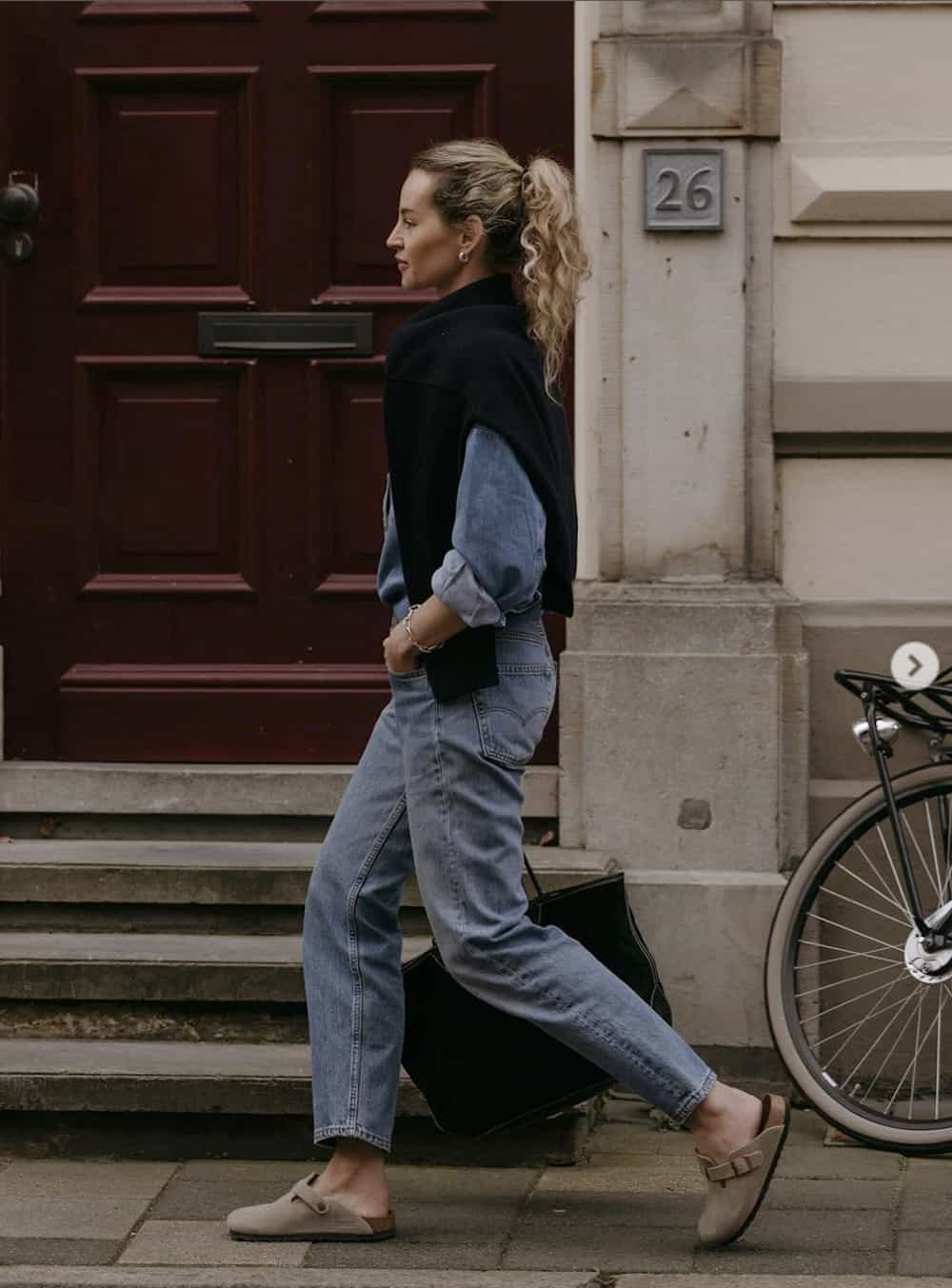 image of a woman walking down the sidewalk in a denim shirt, jeans, and mules with a sweater over her shoulders