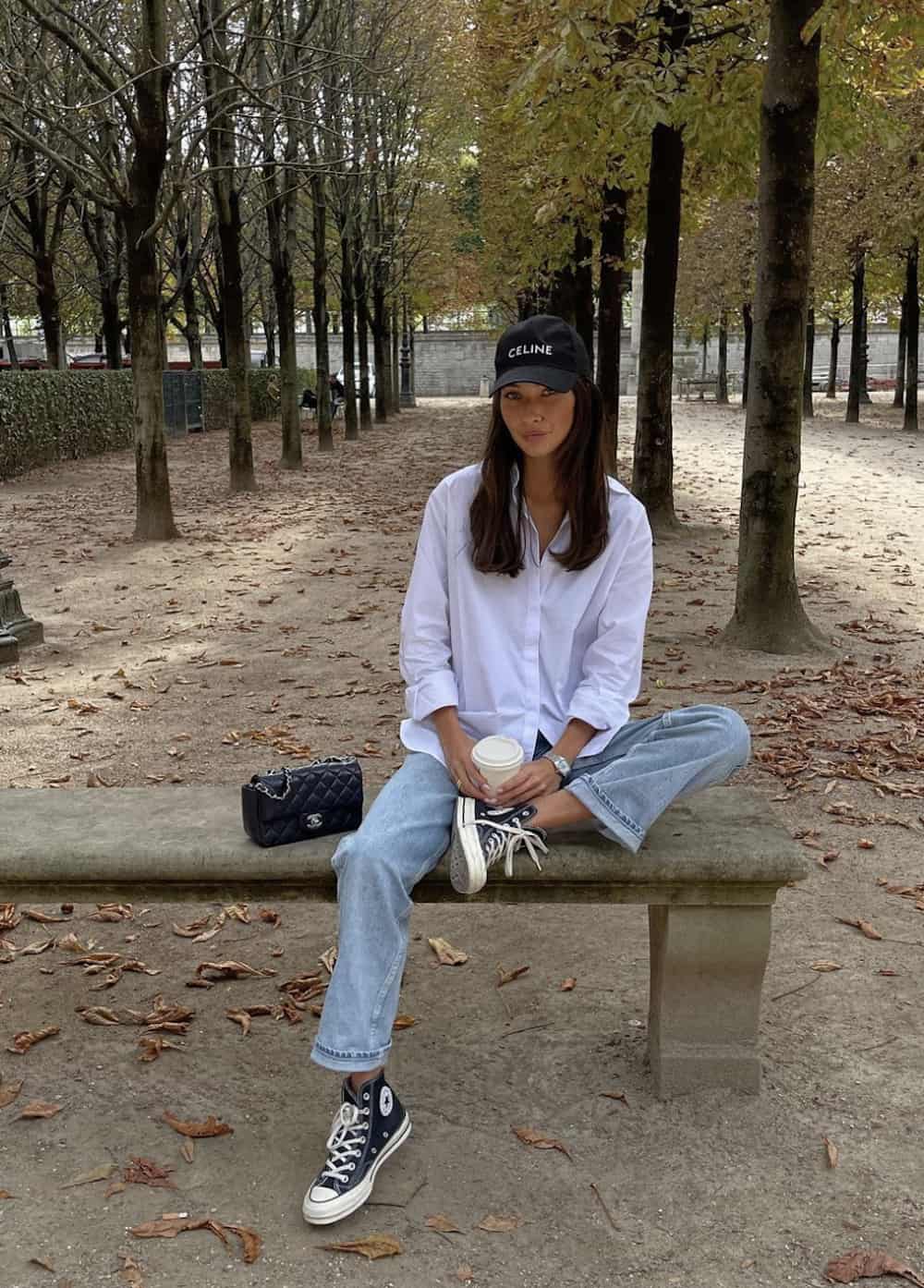 image of a woman sitting on a bench in a park wearing a white button up shirt, jeans, and high top sneakers