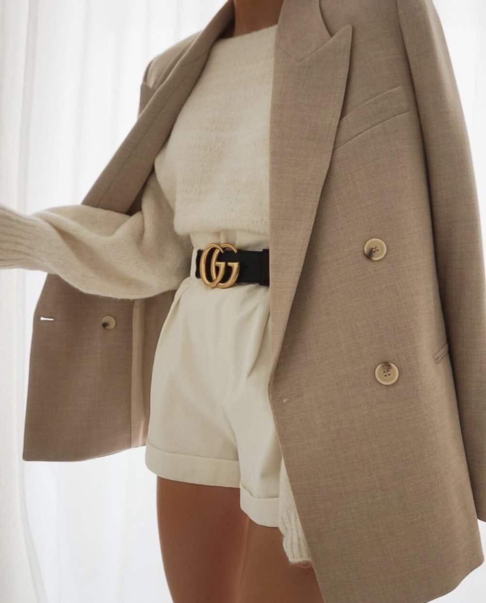 image of a woman from the shoulders down wearing a white sweater and white shorts with a black logo Gucci belt and an oversized beige blazer over her shoulders
