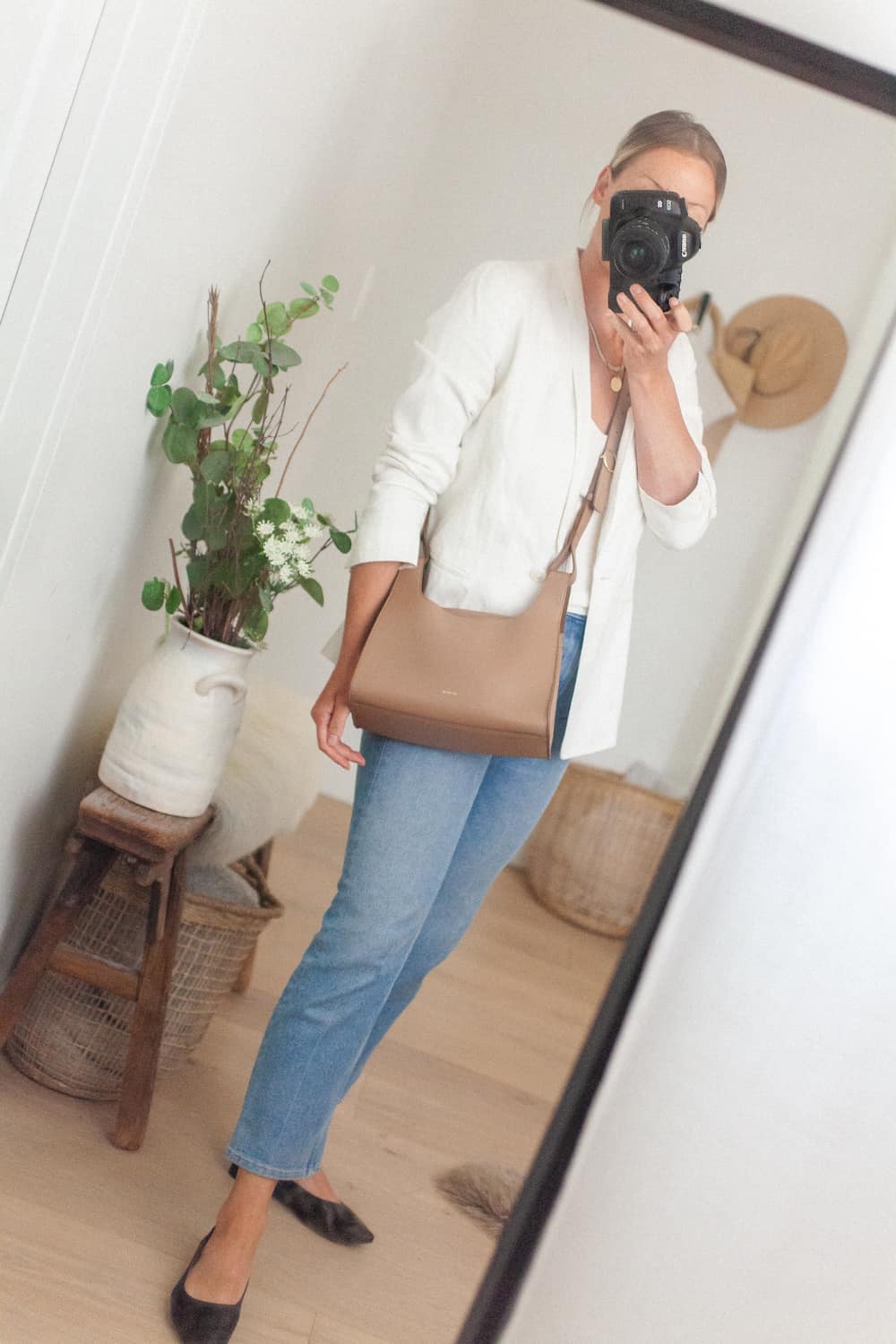 image of a woman wearing a white blazer, blue jeans, black flats and a light brown leather crossbody bag from Cuyana