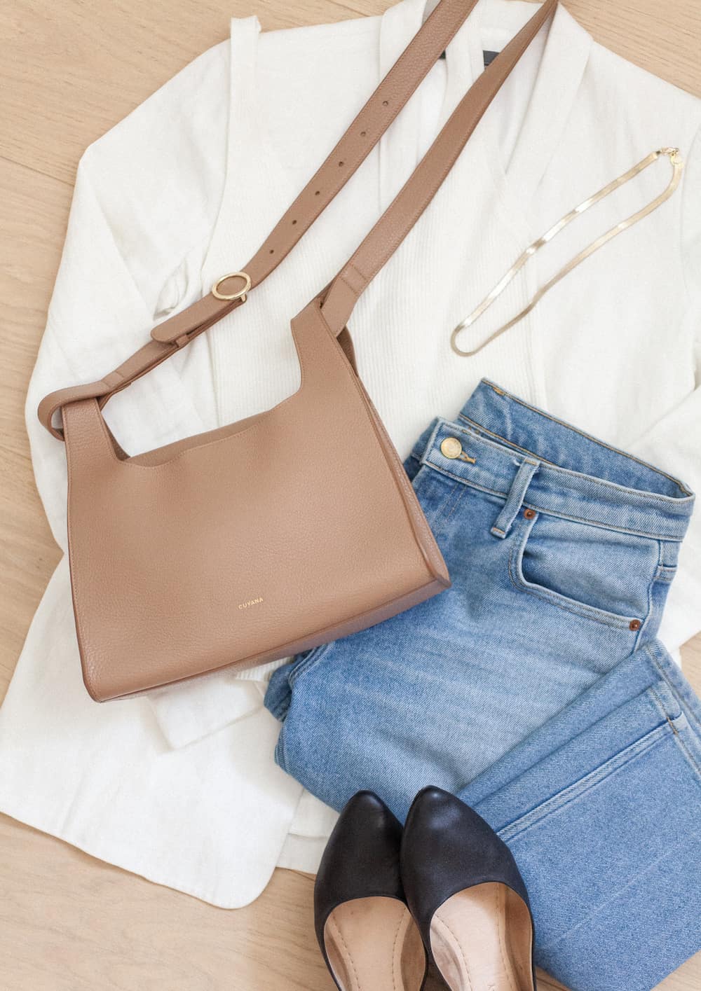 image of n outfit flatlay with a light brown leather Cuyana bag with a white blazer, blue jeans, black flats, and a gold necklace