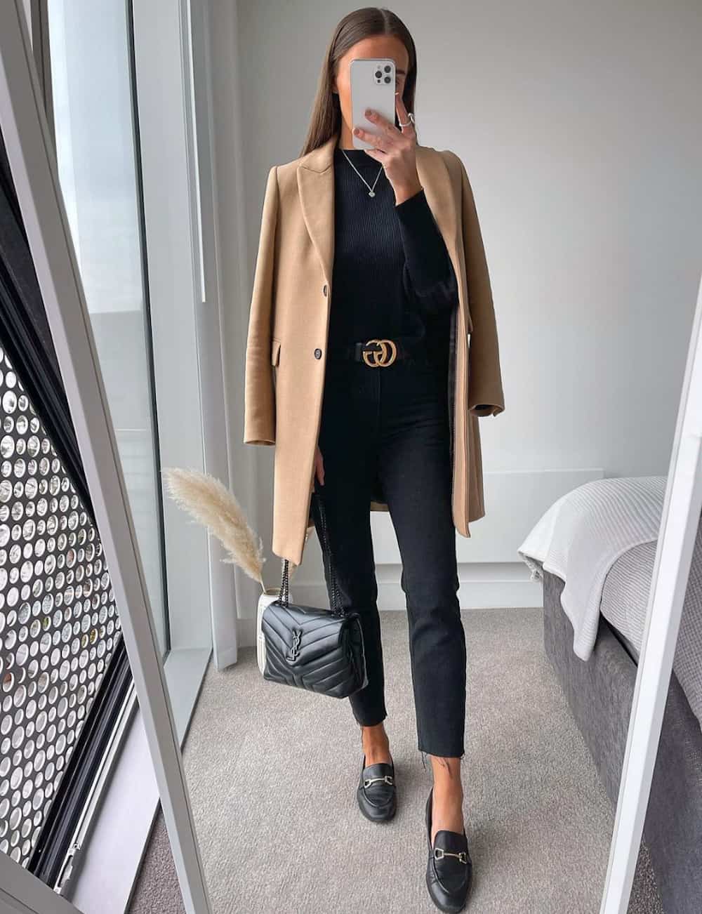 image of a woman wearing a long camel coat with a black sweater, black Gucci belt, and black jeans, and black Gucci loafers