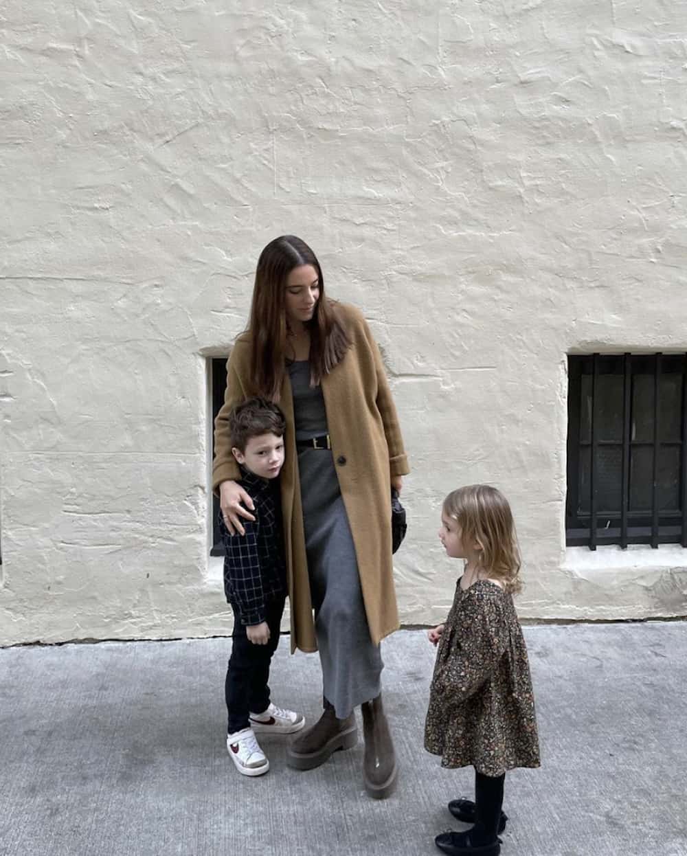 image of a mother with her two children, she is dressed in a long wool camel coat, grey sweater dress, and suede boots