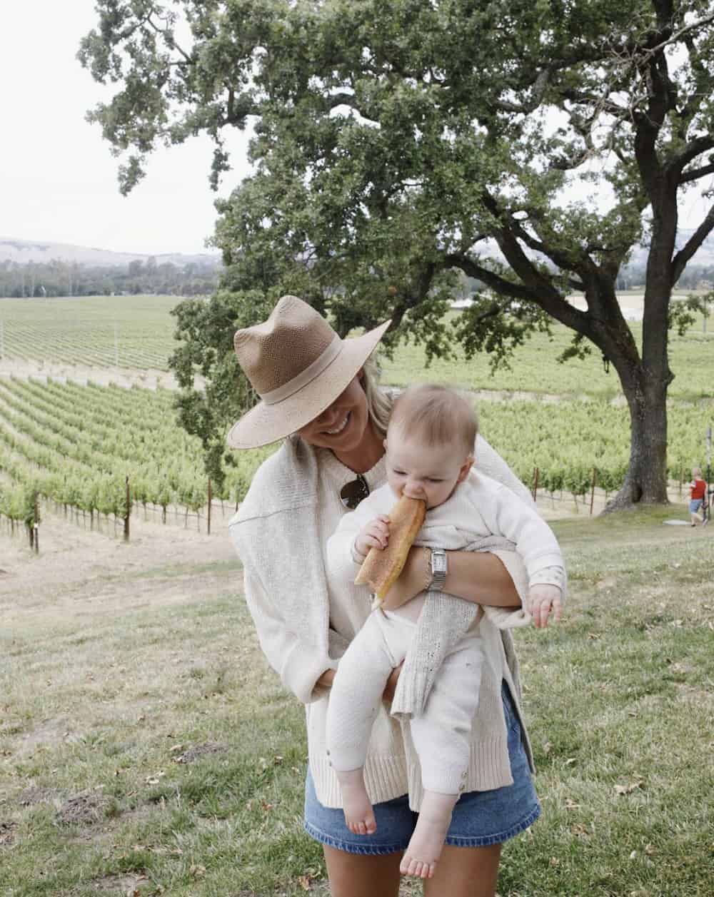 a woman holding a baby, she is a wearing a beige sweater, wool hat, and blue denim shorts