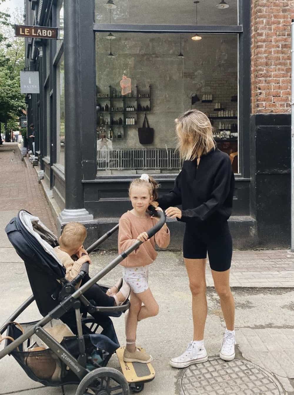 image of a mother pushing a stroller with her two children, wearing a black sweater and black biker shorts with sneakers