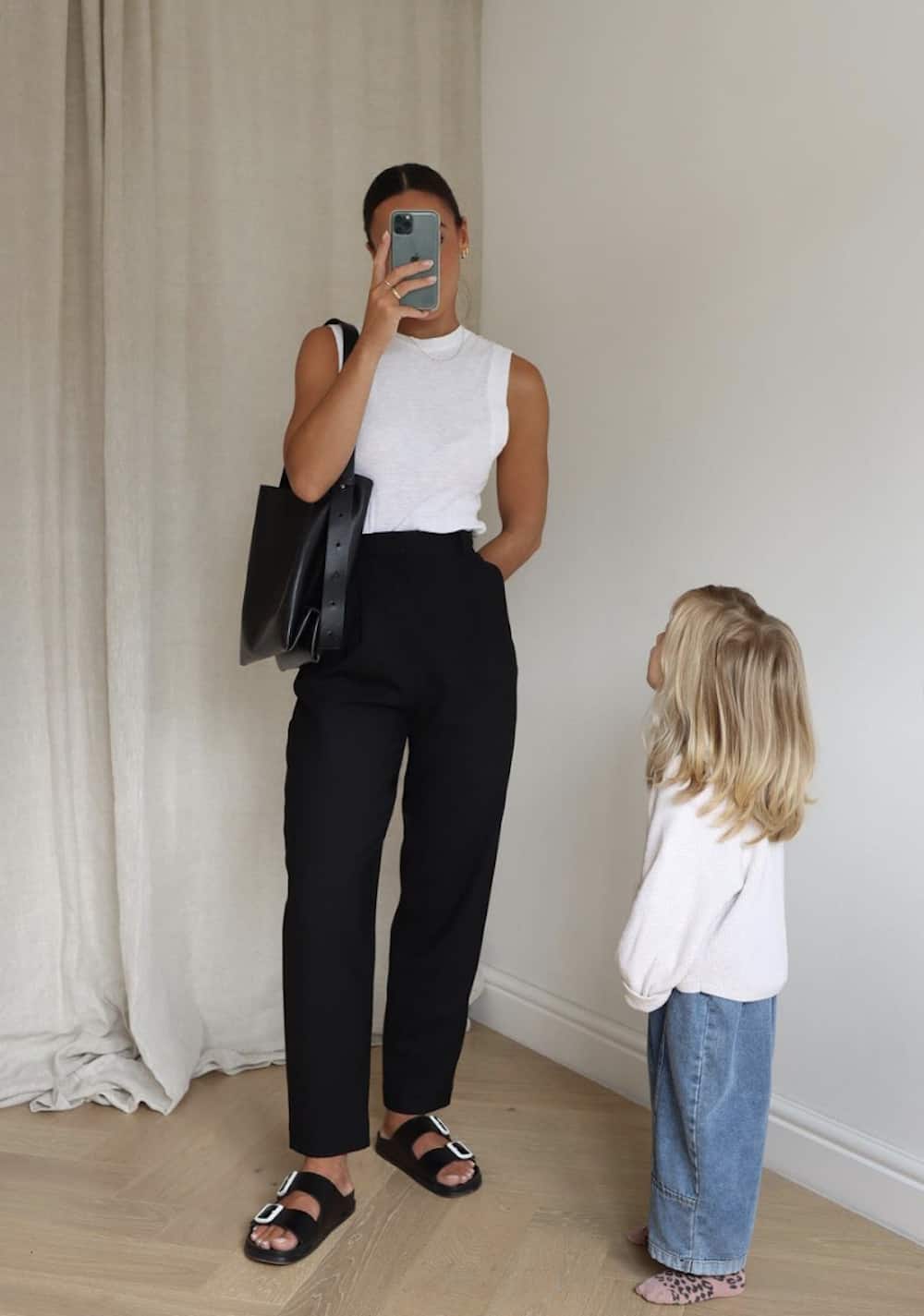 image of a mom and her toddler, wearing a white tank top, black pants, black sandals, and a black tote bag