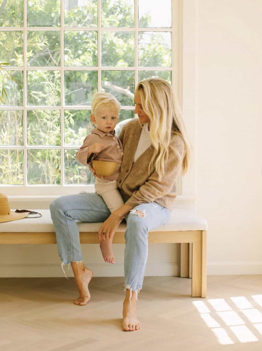 image of a blonde woman sitting on a bench holding her small son, wearing a beige cardigan over a white t-shirt, and ripped blue jeans