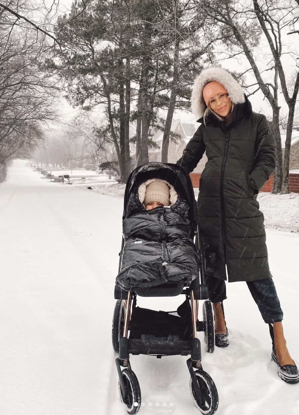 image of a mom with her son in a stroller on a snowy day wearing a long black puffer jacket, and brown lug boots