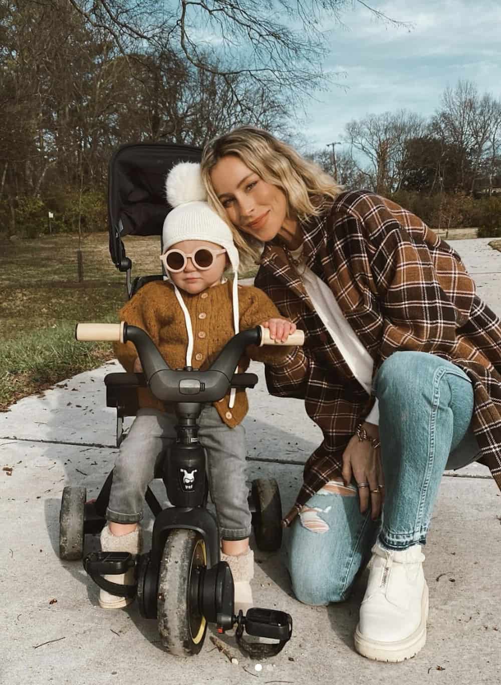 image of a mom leaning over beside her child on a kids bike, wearing a plaid over-shirt, jeans, and white sneakers