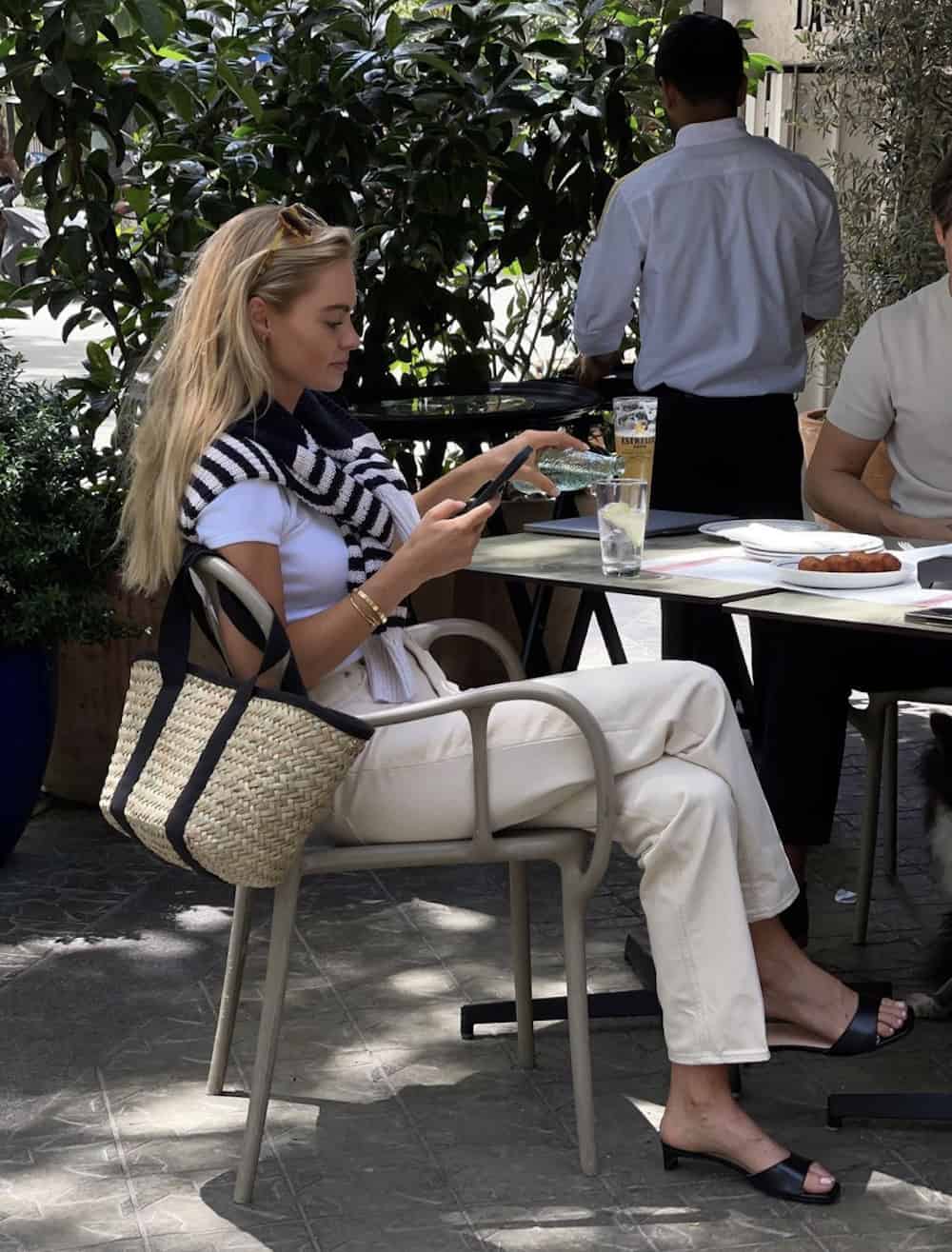 image of a woman sitting at a dining chair on a patio wearing cream jeans, a white t-shirt, and a striped sweater over her shoulders