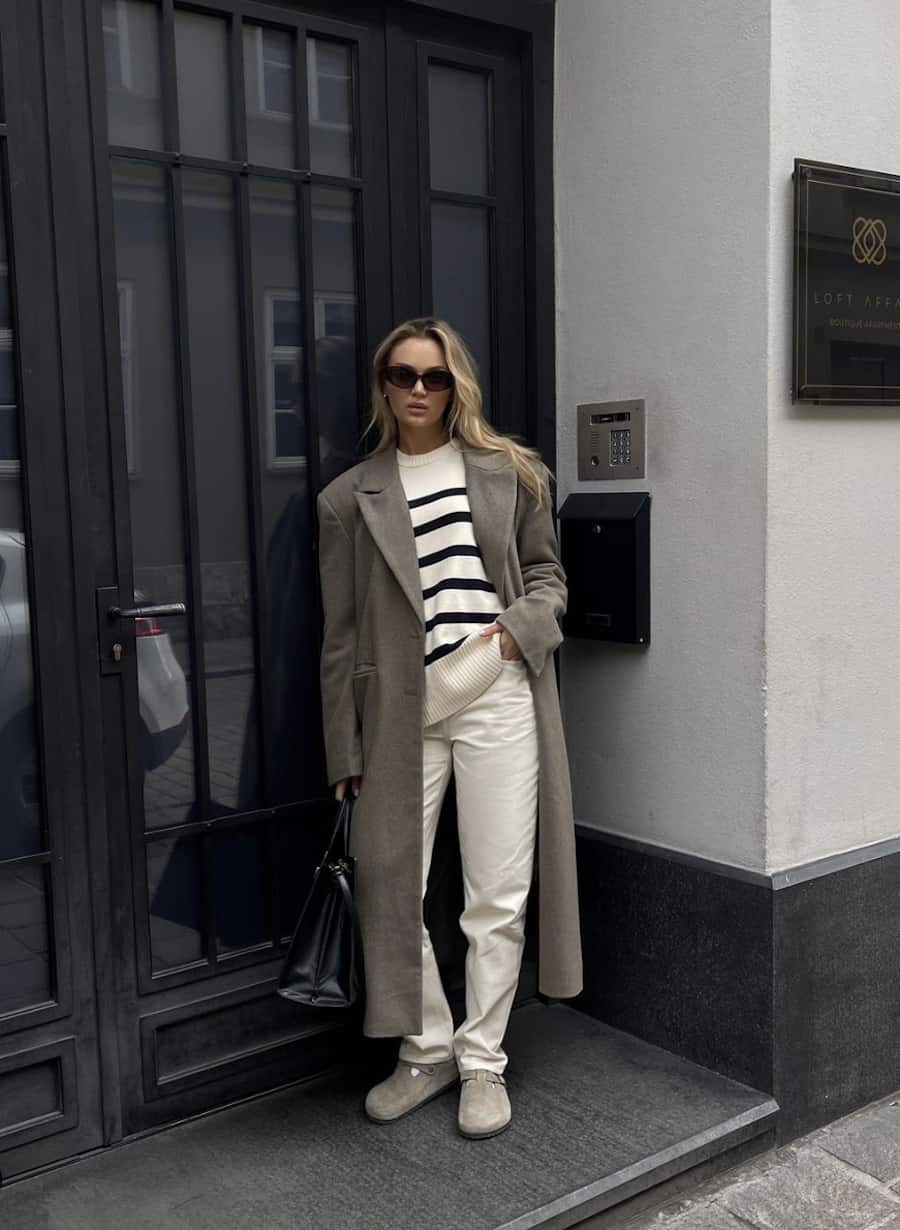 image of a woman in a long wool coat, off white jeans, a striped top, and Birkenstock suede clogs