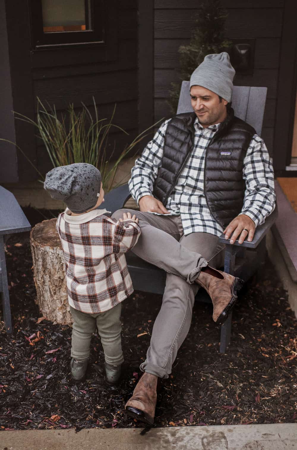 image of a man sitting in a chair wearing grey pants, brown boots, and a plaid shirt, with his son in a plaid jacket and green boots