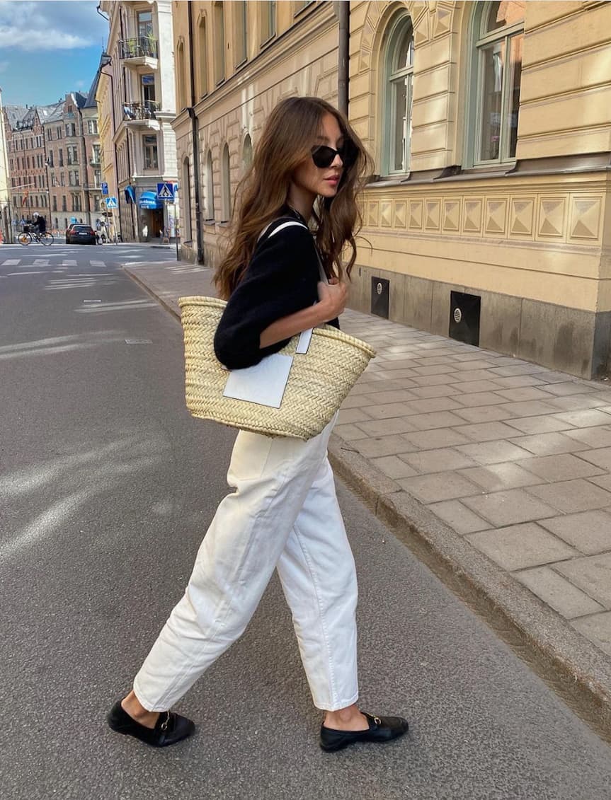 image of a woman walking across the street wearing a black shirt, white jeans, and black shoes with a straw tote bag