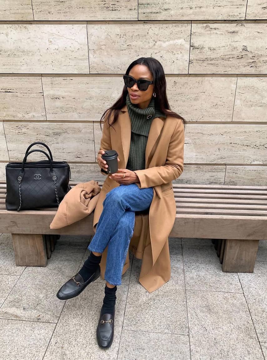 image of a black woman sitting on a bench wearing a stylish outfit with a trench coat, jeans, and black loafers