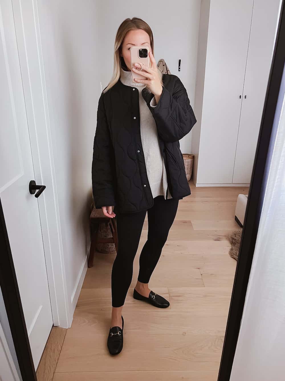 image of a woman wearing an oversized quilted black jacket over a beige turtleneck, black leggings, and black designer loafers
