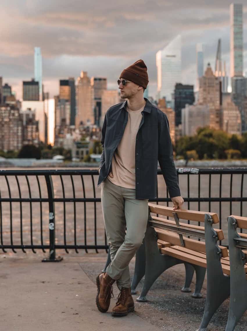 image of a man leaning against a park bench wearing light green chino pants, brown lace-up boots, a green jacket and a beanie