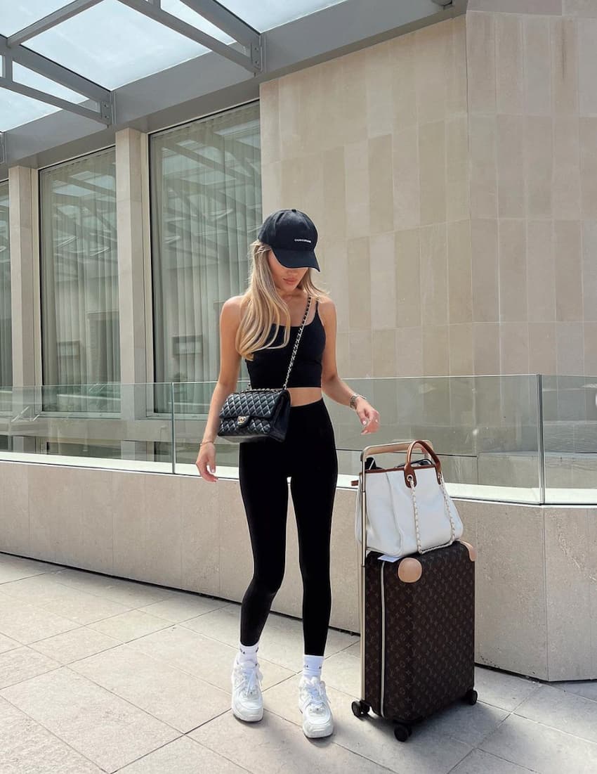 image of a woman standing outside with luggage wearing a crop top, black leggings, a ball cap, and white sneakers