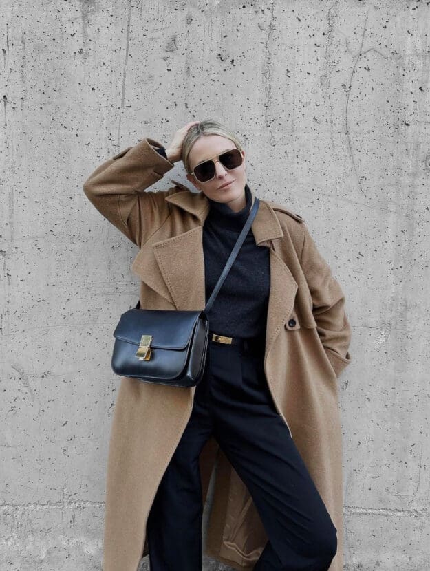 Woman wearing a camel coat over a black sweater and black pants with a black Celine Box bag