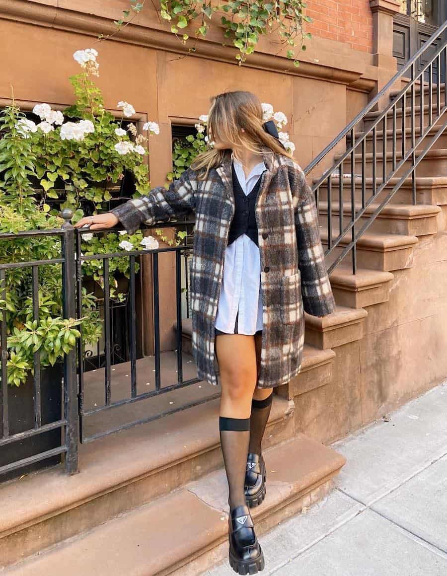 woman wearing a plaid long shirt jacket over a white button down, black mini skirt, with knee high stockings and chunky black loafers