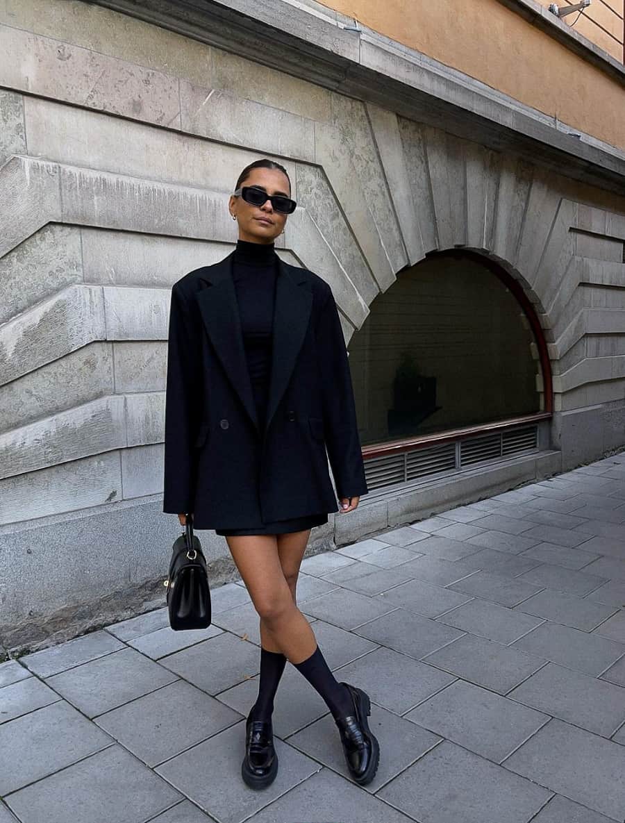 image of a woman wearing an all black outfit with a blazer, turtleneck mini dress and knee-high socks with loafers 