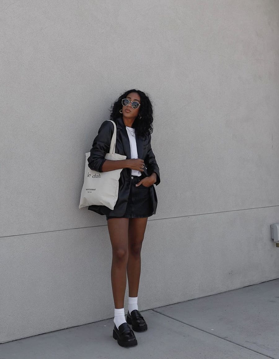 image of a black woman in a black leather blazer, white t-shirt, black denim skirt, white ankle socks, and chunky black flats