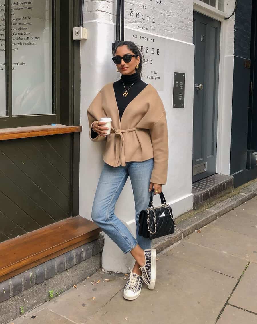 woman wearing a black turtleneck, camel wrap coat, blue jeans, and sneakers holding a coffee cup