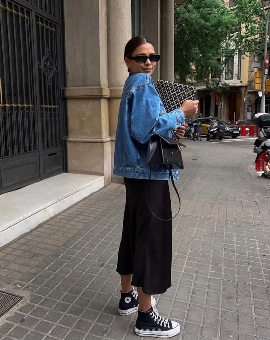 woman wearing an oversized denim jacket, black midi skirt, and converse high-top sneakers