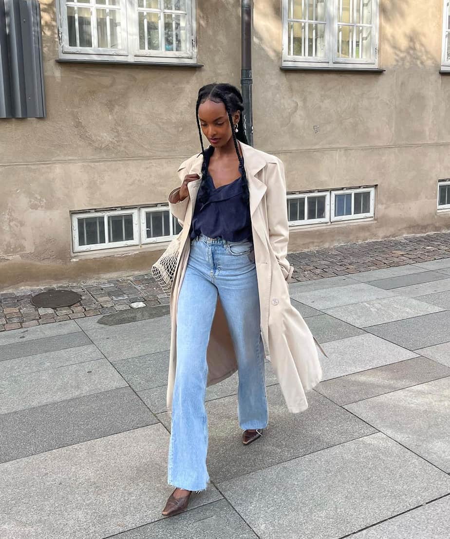 black woman wearing a long tan trench coat, blue jeans, a navy camisole top, and heels