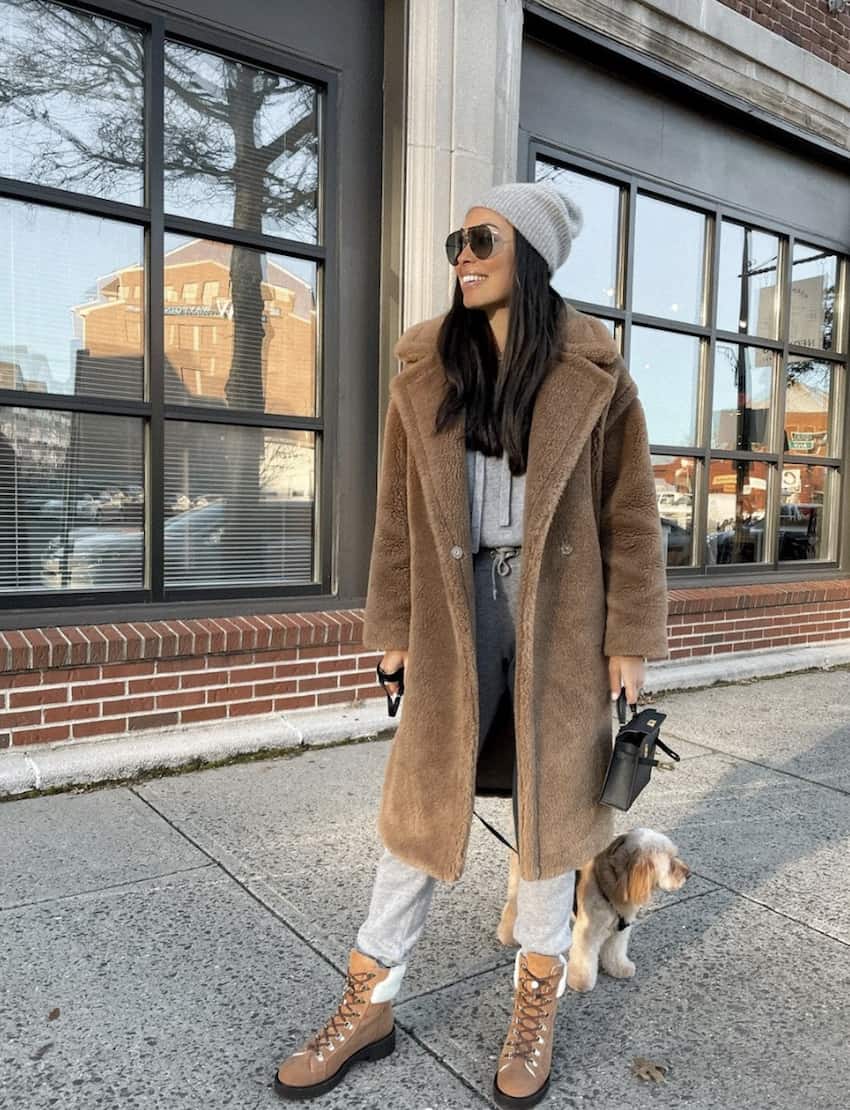 woman wearing a long faux fur coat, cozy pants, boots, and a beanie