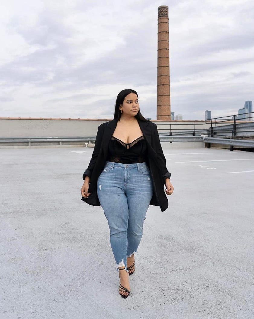 plus-size woman wearing a black lace bodysuit with skinny jeans, heels, and a black blazer