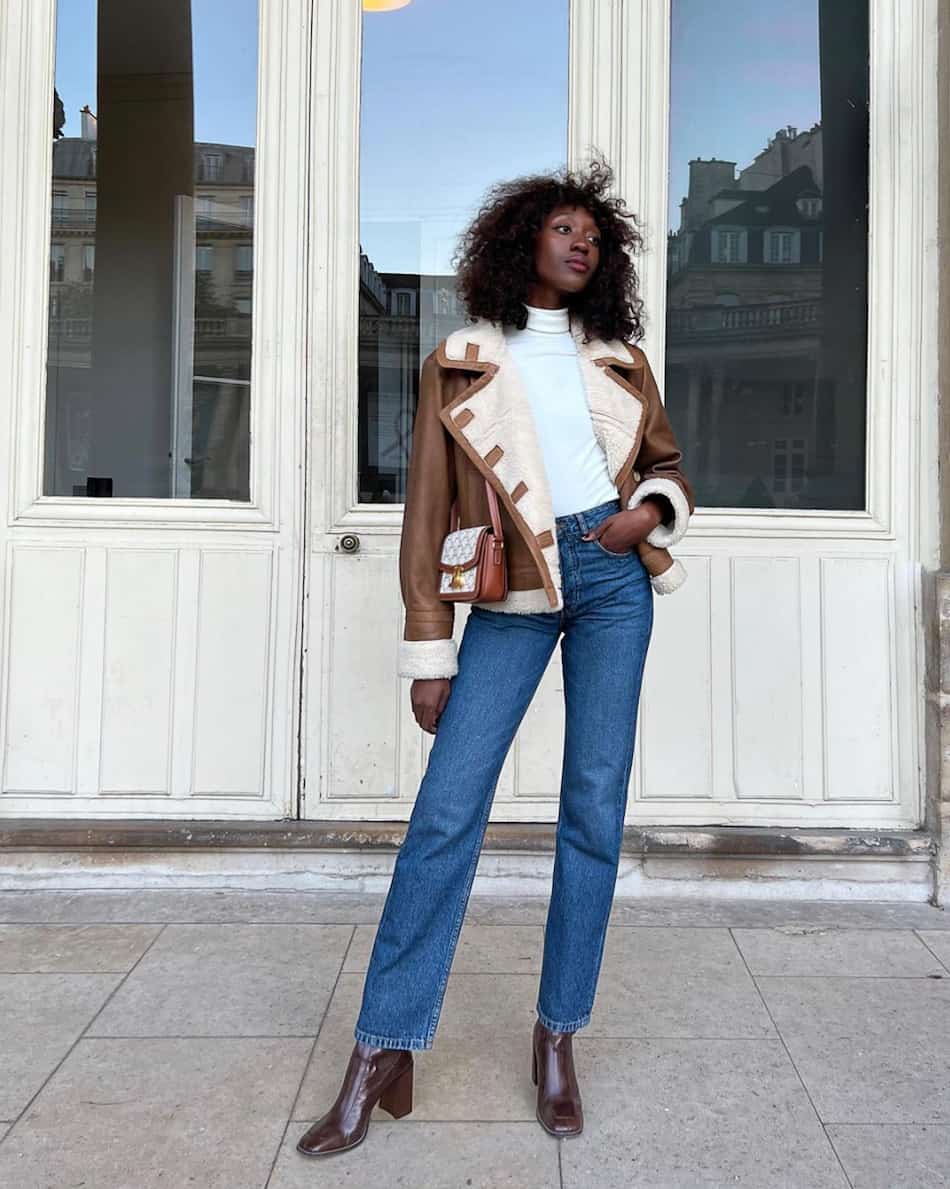 black woman wearing a white turtleneck bodysuit under a shearling leather jacket with blue jeans and brown boots