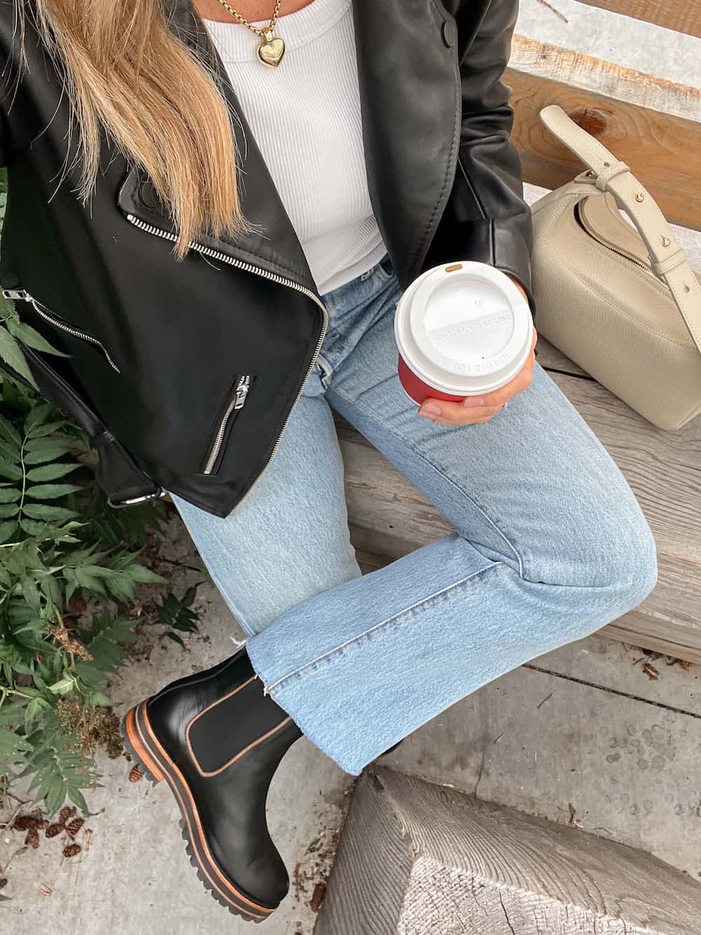overhead view of a woman wearing a black leather coat with jeans and black chelsea boots holding a coffee