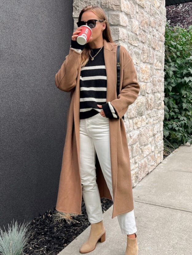 woman wearing a long camel coat over a black striped sweater with off white jeans and suede tan boots drinking a coffee