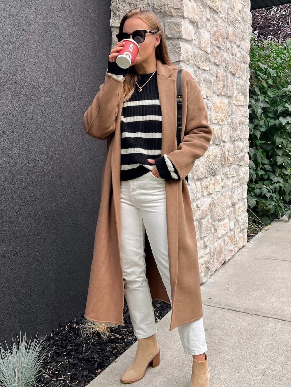 woman wearing a long camel coat over a black striped sweater with off white jeans and suede tan boots drinking a coffee