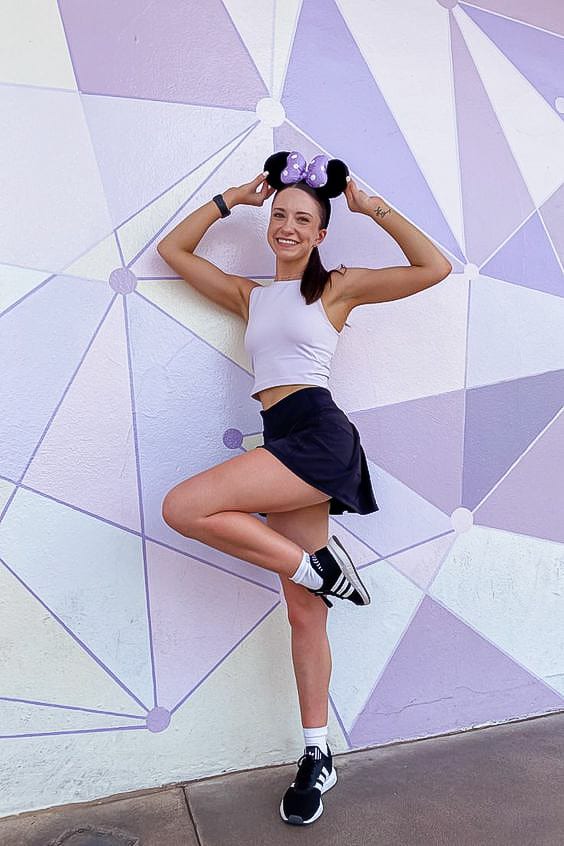 woman standing in front of a purple wall wearing a purple tank, black skirt, black sneakers, and black and purple Minnie Mouse ears
