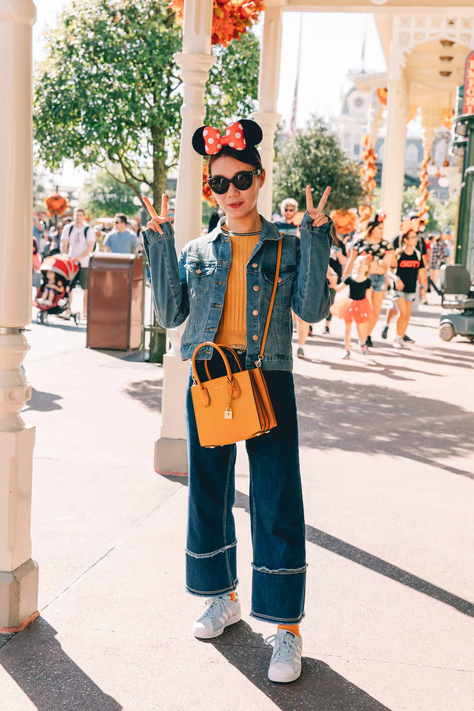 woman wearing a jean jacket, orange sweater, blue jeans, and sneakers with Minnie Mouse ears at Disneyland