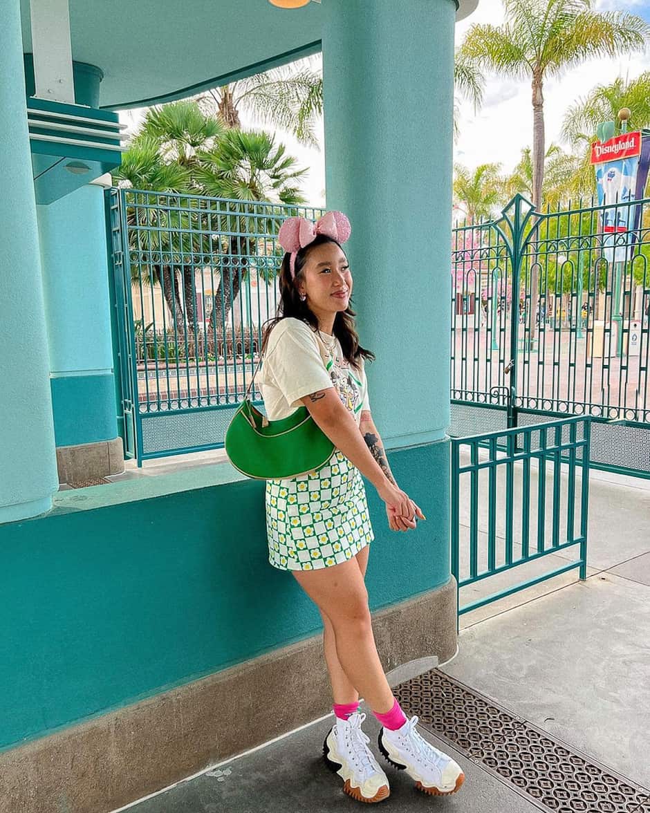 woman at Disneyland wearing a graphic t-shirt, green floral mini skirt, sneakers, and pink glitter Minnie Mouse ears