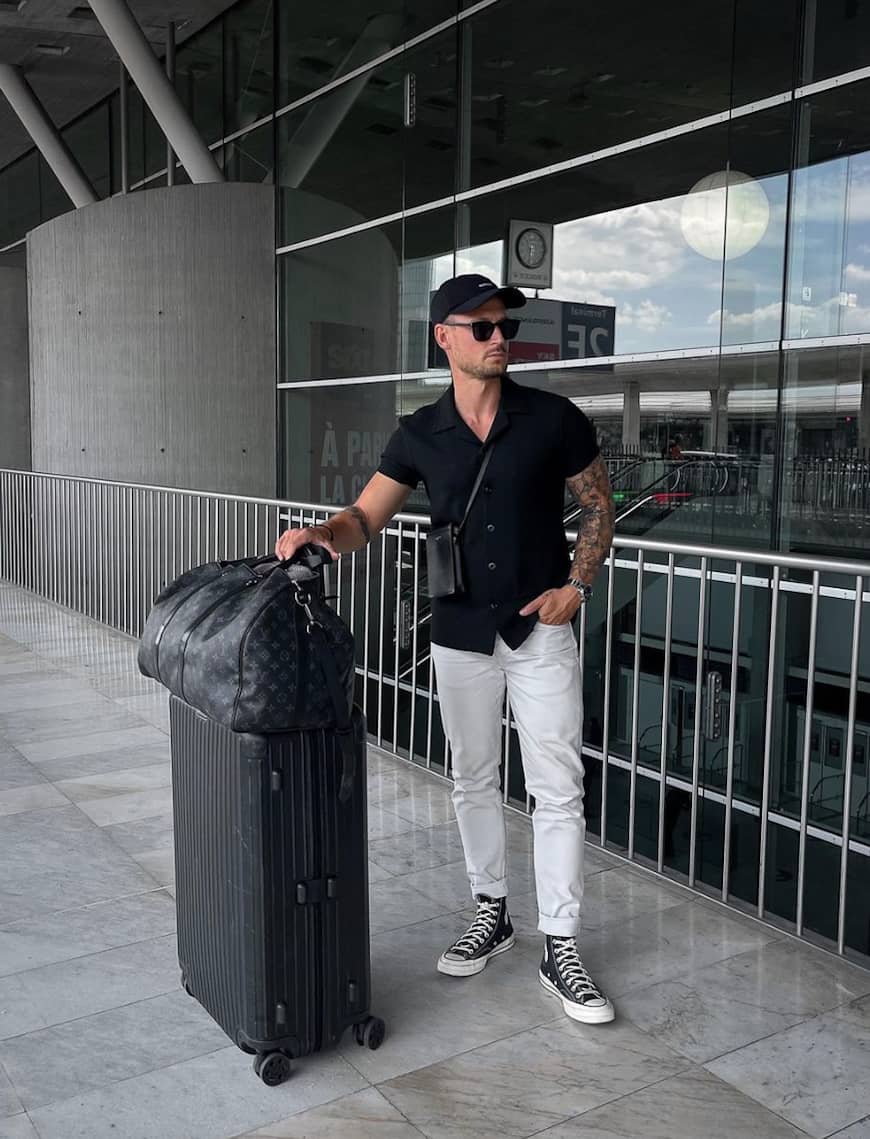 man with luggage wearing a black button up shirt and chino pants with sneakers