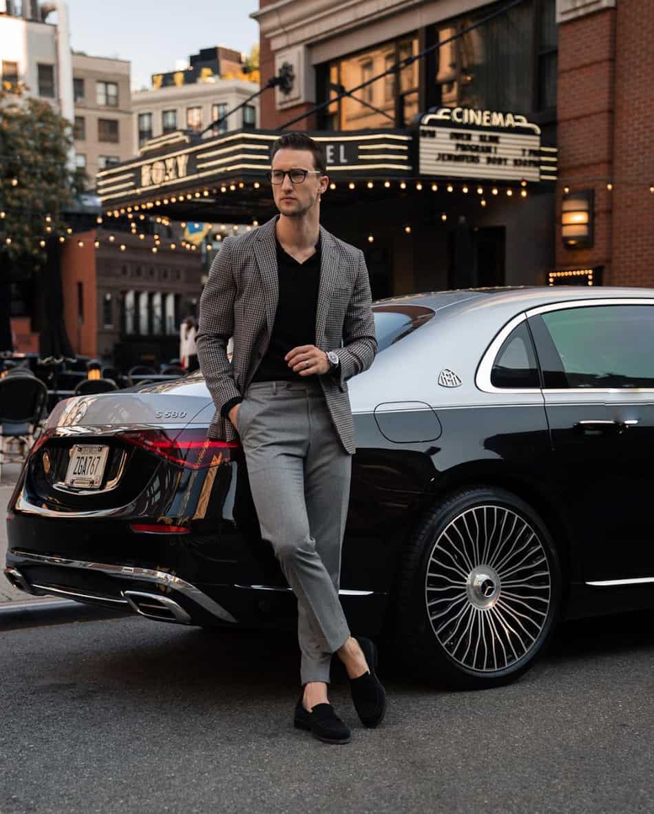 stylish man leaning against a car wearing a blazer, grey pants, a black shirt, and dress shoes