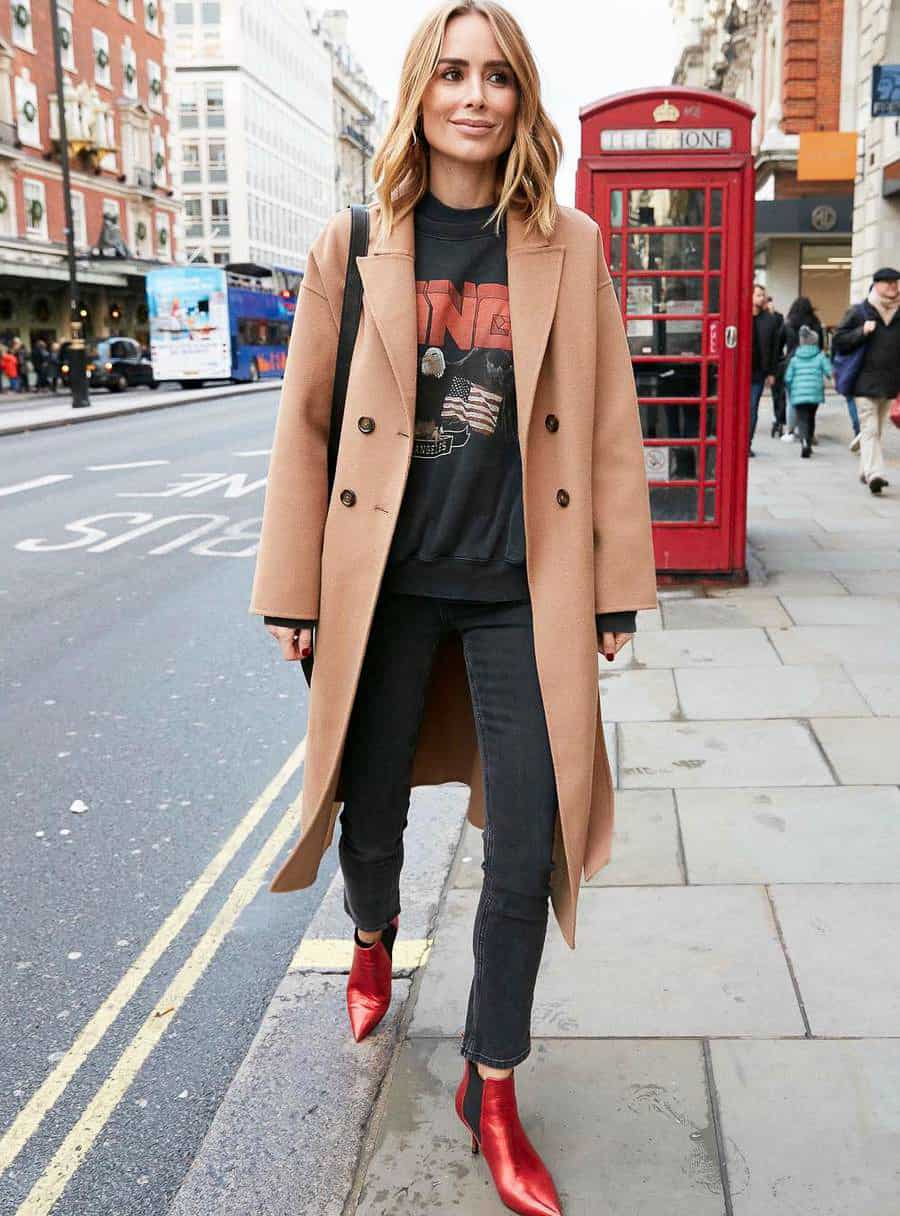 woman wearing a long camel coat over a graphic sweatshirt with black jeans and shiny red leather ankle boots