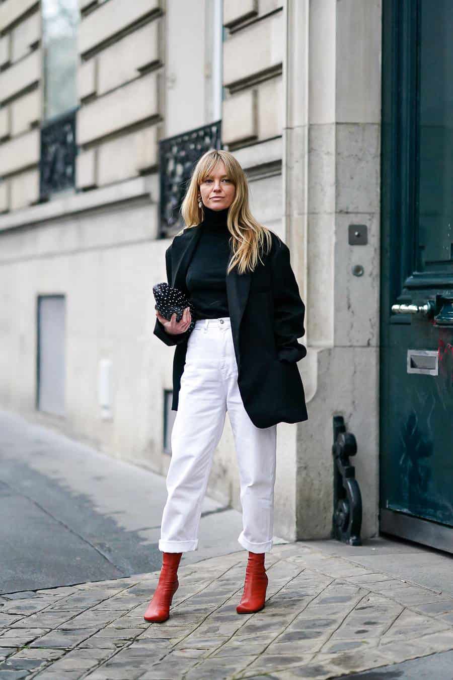 Jeanette Madsen wears earrings, a black turtleneck pullover, a black oversized blazer jacket, white pants, red leather boots, a black bag with polka dots